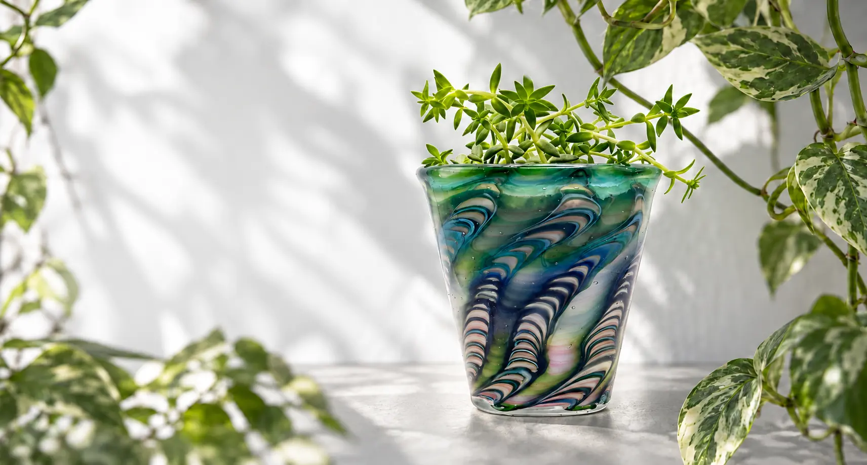 A blue-green glass planter with feathered bands of color and a branching succulent, surrounded by leafy vines
